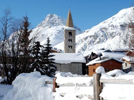 Clocher de pierre de Val-d'Isère et toits enneigés, montagnes au fond