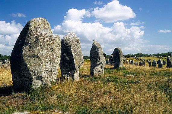 Une file de menhirs couverts de lichen dans l'herbe sèche, sous un ciel bleu à gros nuages blancs, dans les alignements de Carnac