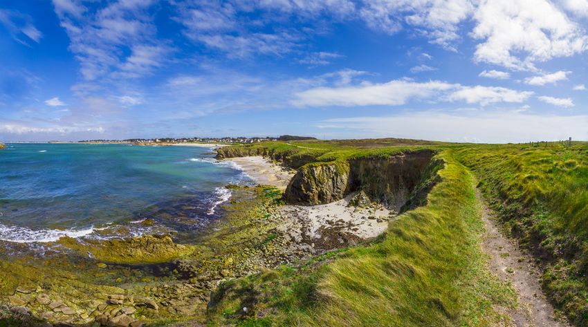 Sentier côtier herbeux au-dessus d'une crique de sable et de rochers, mer turquoise et ciel bleu, sur le littoral du Morbihan