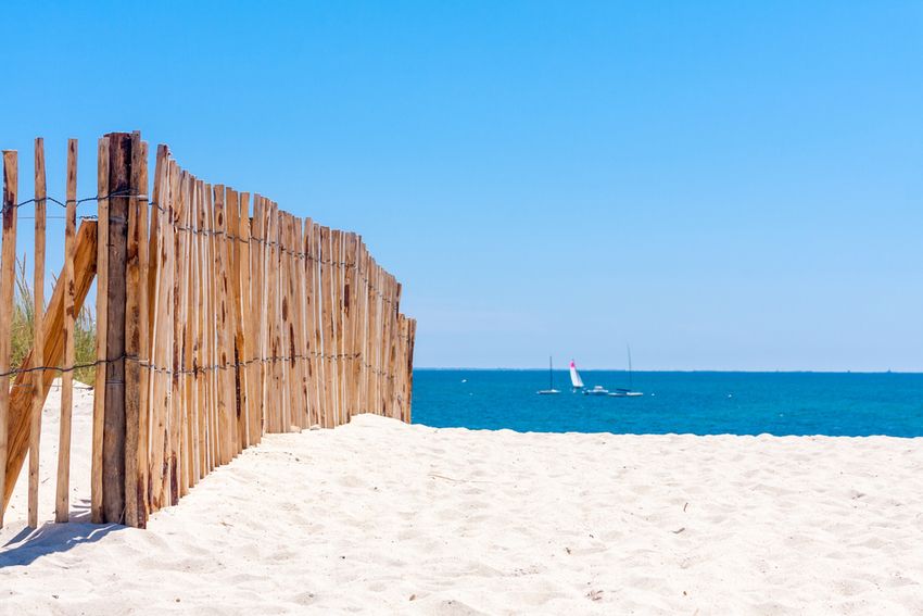 Sable blanc, ganivelles de bois et voiliers sur une mer bleu profond, sur une plage de Carnac en été
