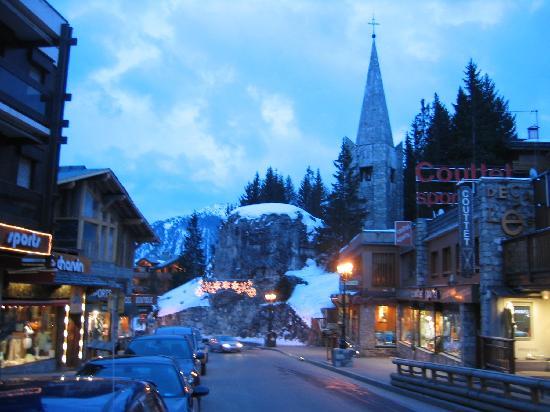 Rue commerçante de Courchevel au crépuscule, clocher de pierre et rocher enneigé illuminé
