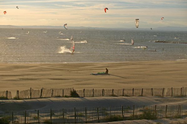 Kitesurfeurs et véliplanchiste sur la mer au coucher du soleil, vus depuis une plage de sable bordée de ganivelles, à Port-Camargue