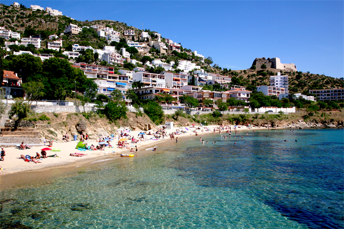Petite plage de sable et eau turquoise au pied de maisons étagées, avec la forteresse de la Trinitat au sommet de la colline