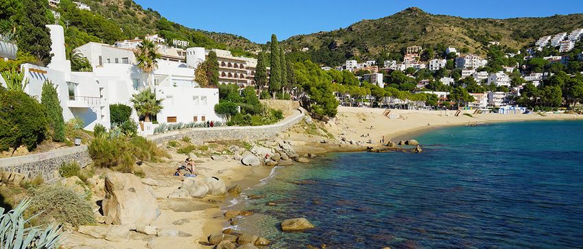 Petite plage de sable en anse, bordée de rochers, de pins et de maisons blanches à flanc de colline, sur la côte est de Roses