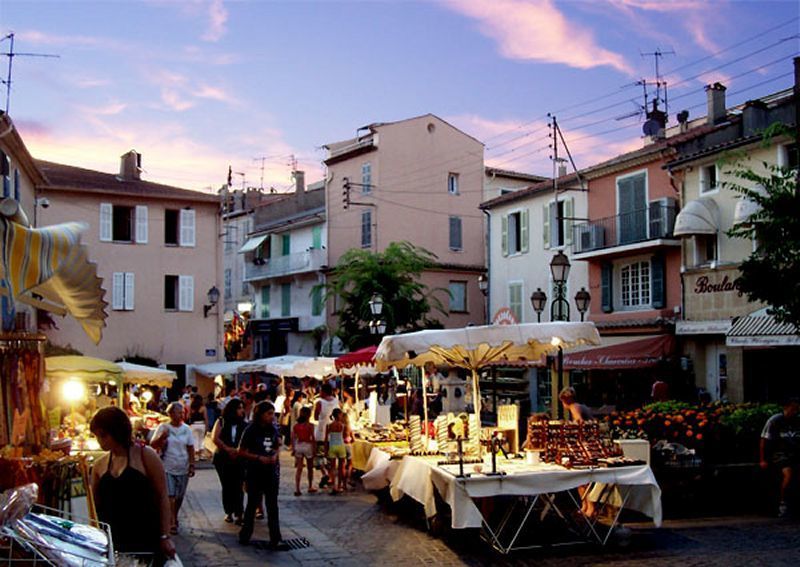 Étals d'un marché provençal du soir sur une place, entre façades colorées et réverbères, sous un ciel rose de fin de journée